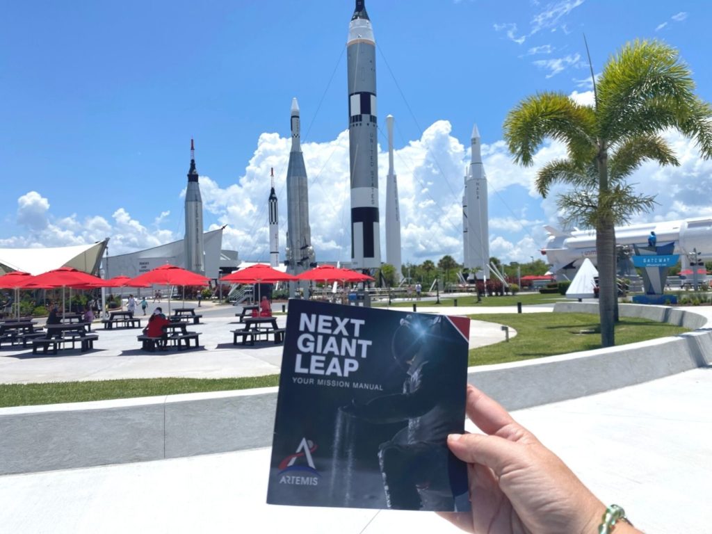 Dani Meyering holds the Artemis Mission Manual Scavenger Hunt at Kennedy Space Center Visitors Complex in front of the camera with the rocket garden in the background