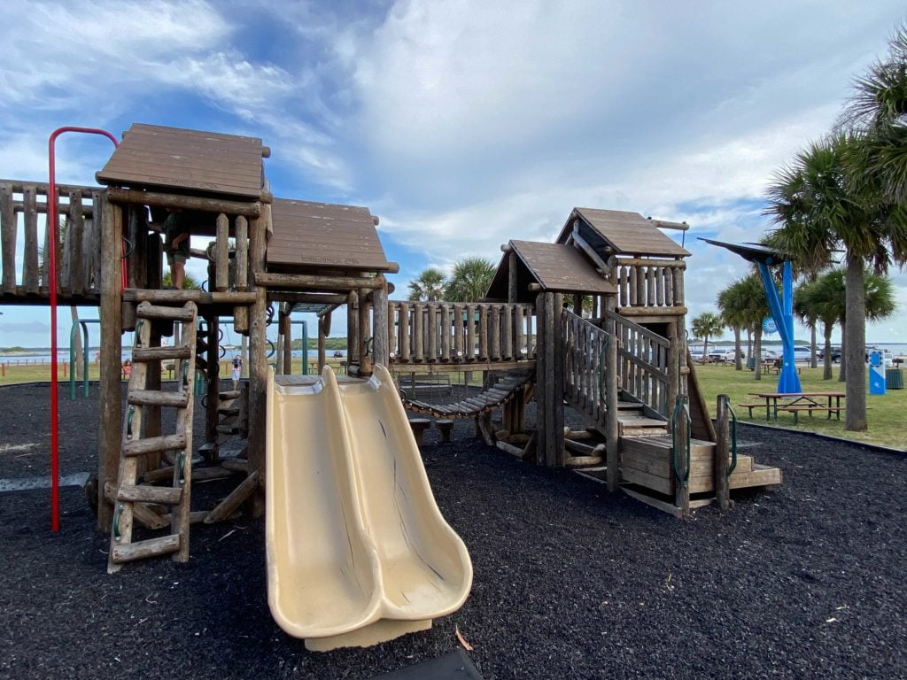 Jetty Park Playground is wooden with multiple slides, climbing structures, and shredded black rubber as padding around the playground. 