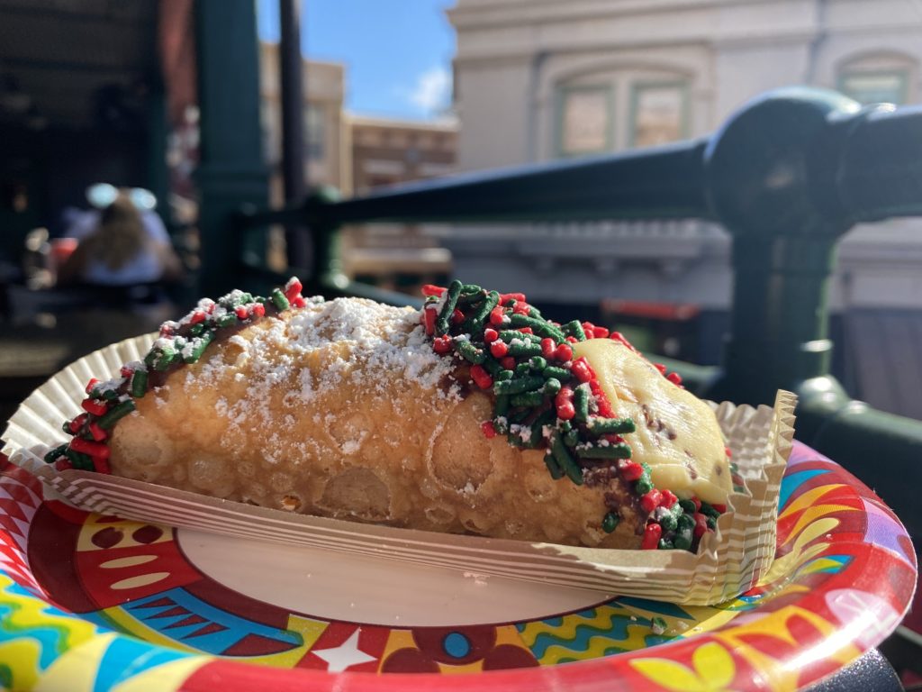 Butterscotch Caramel Cheesecake Cannoli at PizzeRizzo, the cannoli is in the foreground on a Disney World paper plate with chocolate dipped on each end of the cannoli and red and green sprinkles, and buildings that are part of the Muppets courtyard in the background and a bit of blue sky