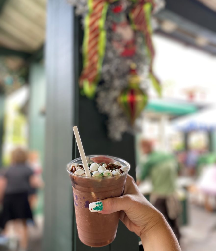 Salted Caramel Frozen Hot Chocolate at Most Quick Service Locations at Hollywood Studios is in the foreground held up by the photographer, with her hand in the foreground holding up the cup of frozen hot chocolate, and silver tinsel holiday decorations in the blurred background with bright green and red ornaments