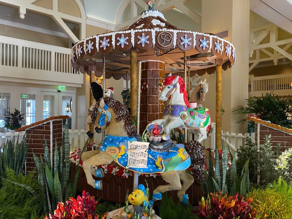 2021 Gingerbread Carousel at Disney's Beach Club Resort - The Little Mermaid theme - Two carousel horses made of gingerbread and chocolate are at the front of this photo. The horse in the foreground is decorated with bright, light blue and green and yellow colors with the characters Flounder and Sebastian. The horse just behind it is themed after Ariel, with red mane and purple and sea green colors.