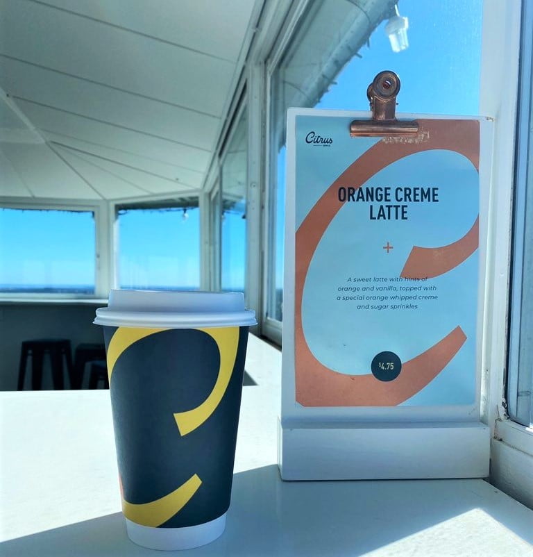 A paper coffee cup with a dark background and a curvy letter C from Citrus Coffee at Florida Citrus Tower is in the foreground next to a small sign advertising the Orange Creme Latte. The observation deck with a clear blue sky are in the background