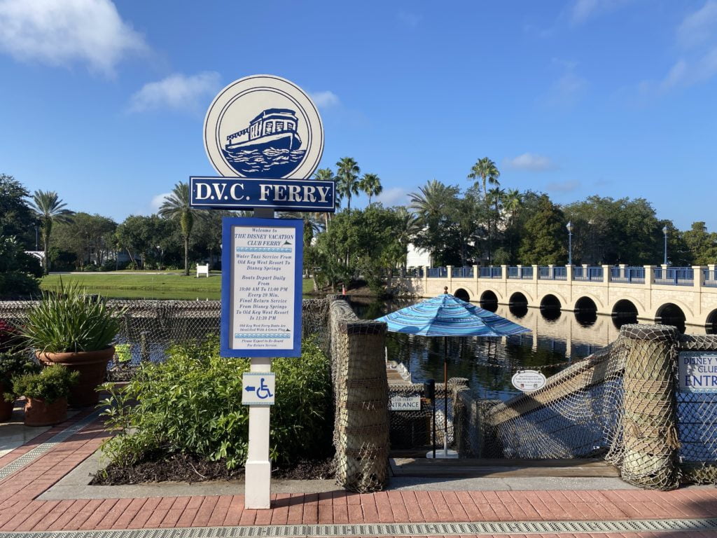 Disney's Old Key West Resort - Home to Olivia's Cafe and a Ferry Boat - This photo shows the DVC Ferry sign in the foreground with beige and dark blue colors, and the boat dock in the background with a blue umbrella on the dock and a roadway bridge in the very back of the photo and blue skies above.