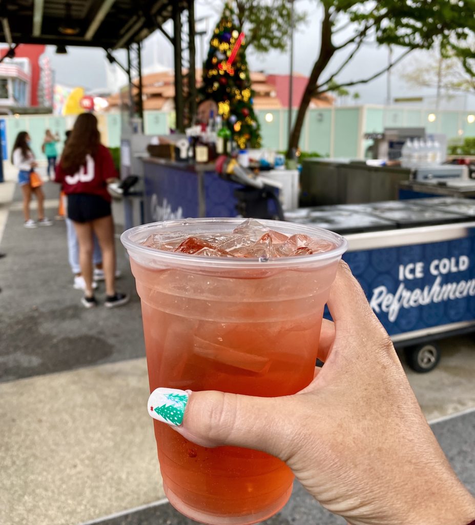 The Fireside Cider at Disney Springs Outdoor Carts has a pale red color and it is served in a plastic clear cup with ice. This picture was taken with the photographer holding the drink in her hand, in the foregrounf, with the food and beverage cart and a Christmas tree in the background