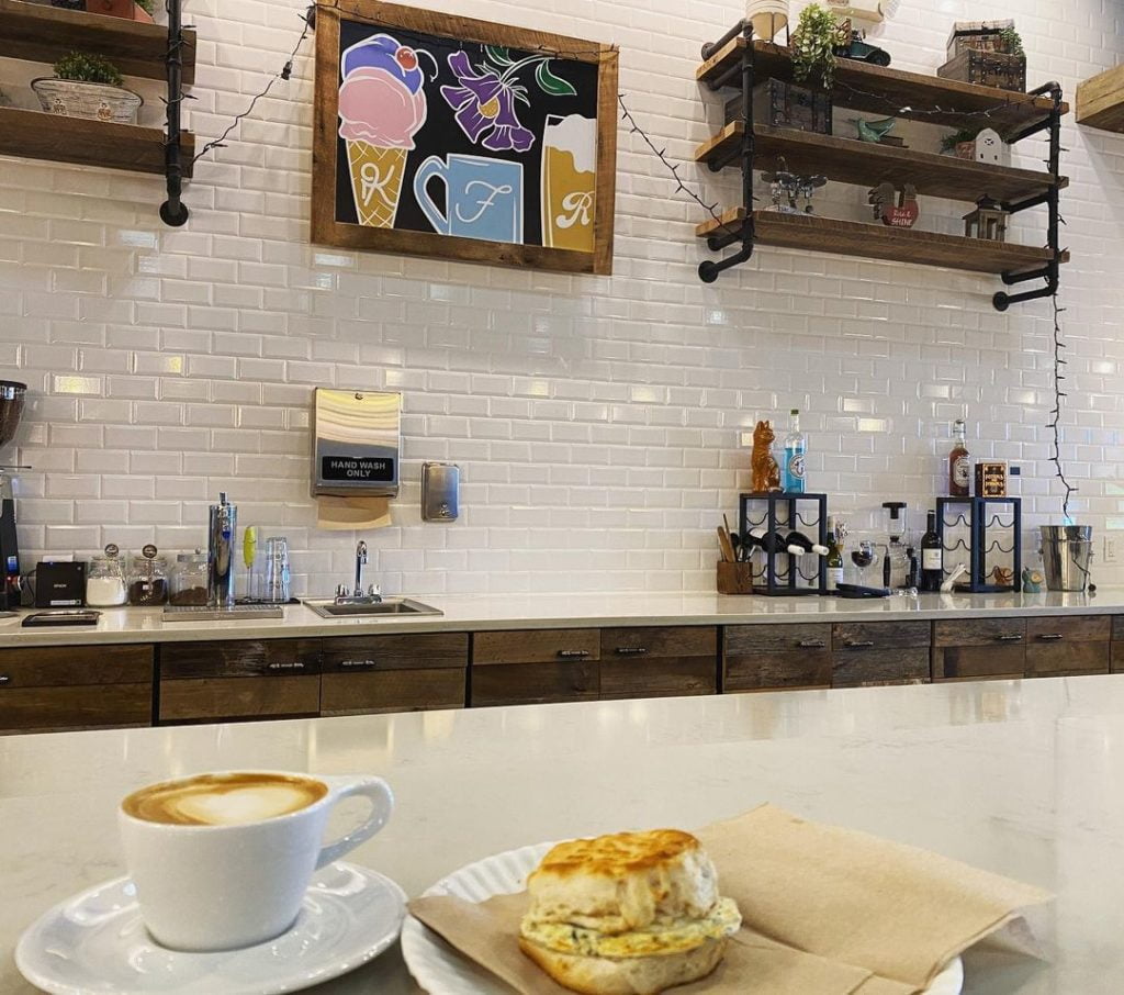 Foxtail Howell branch location with bar seating - ceramic mug with latte sits next to a vegetable and egg sandwich on a ceramic plate, set on the white counter with a farmhouse style wall and a chalkboard style piece of art that shows the logos of Kelly's Homemade Ice Cream Foxtail Coffee and Ravenous Pig in the background
