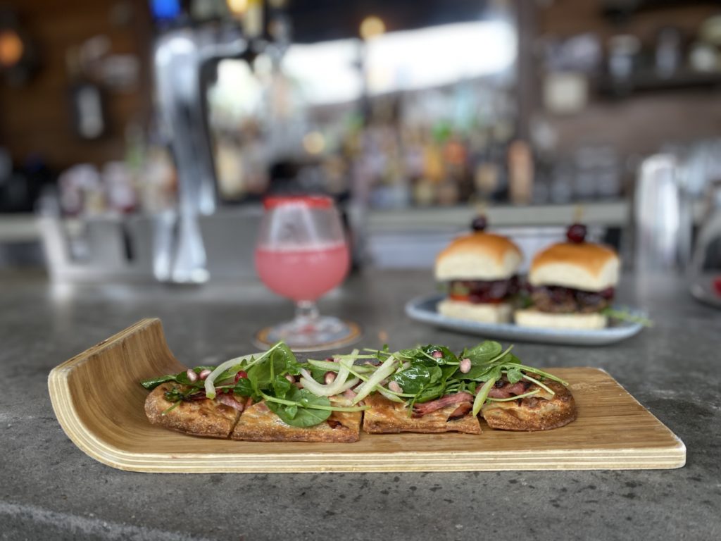 Ham and Brie Flatbread is served on a wooden tray that resembles a sled, and a red colored cocktail and a pair of venison sliders are placed behind the flatbread at Jock Lindsey's Hangar Bar