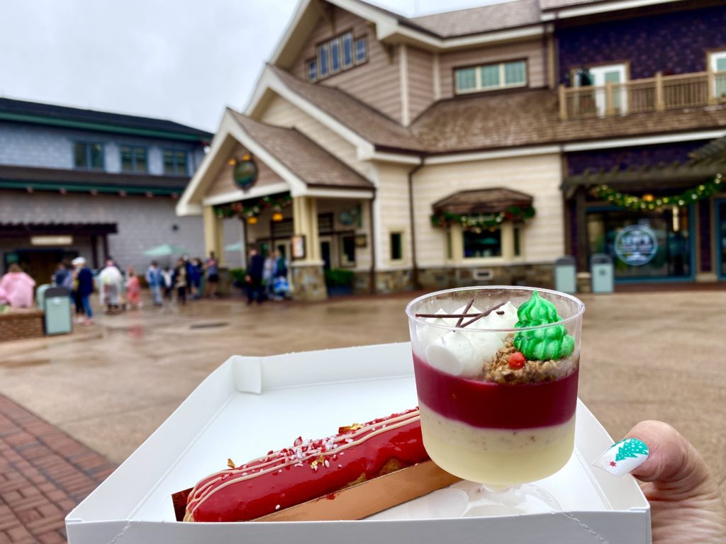The White Chocolate Peppermint Eclair at Amorette's Patisserie are in the foreground with festive red toppings. The photographer is holding a small tray with these sweets and Amorette's Patesserie is in the background and has a cream and brown colored exterior. 