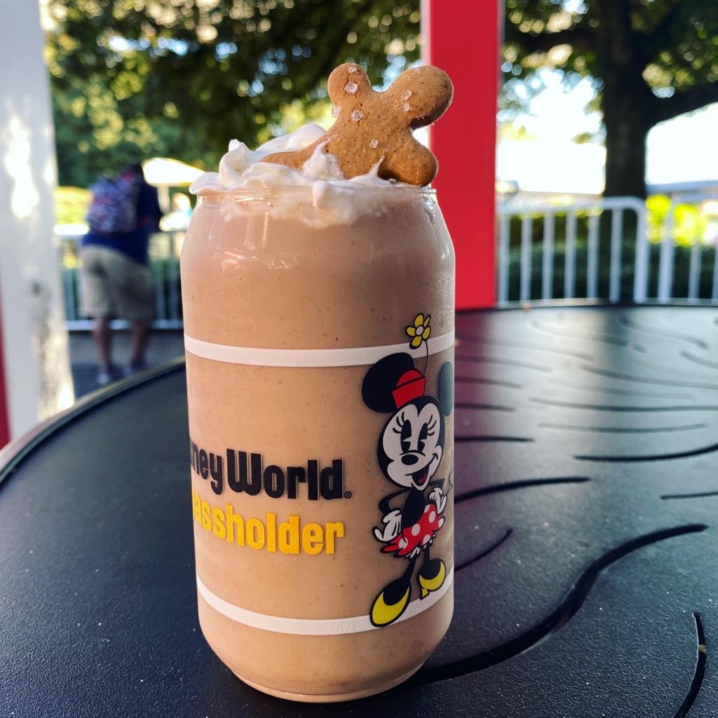 Gingerbread Milkshake at The Donut Shop EPCOT Festival of the Holidays is served in a clear plastic cup with Minnie Mouse and the words Walt Disney World Passholder in black and yellow, wrapped around the middle of the cup. The milkshake within is a light brown color and is topped with whipped cream and a mini gingerbread man cookie