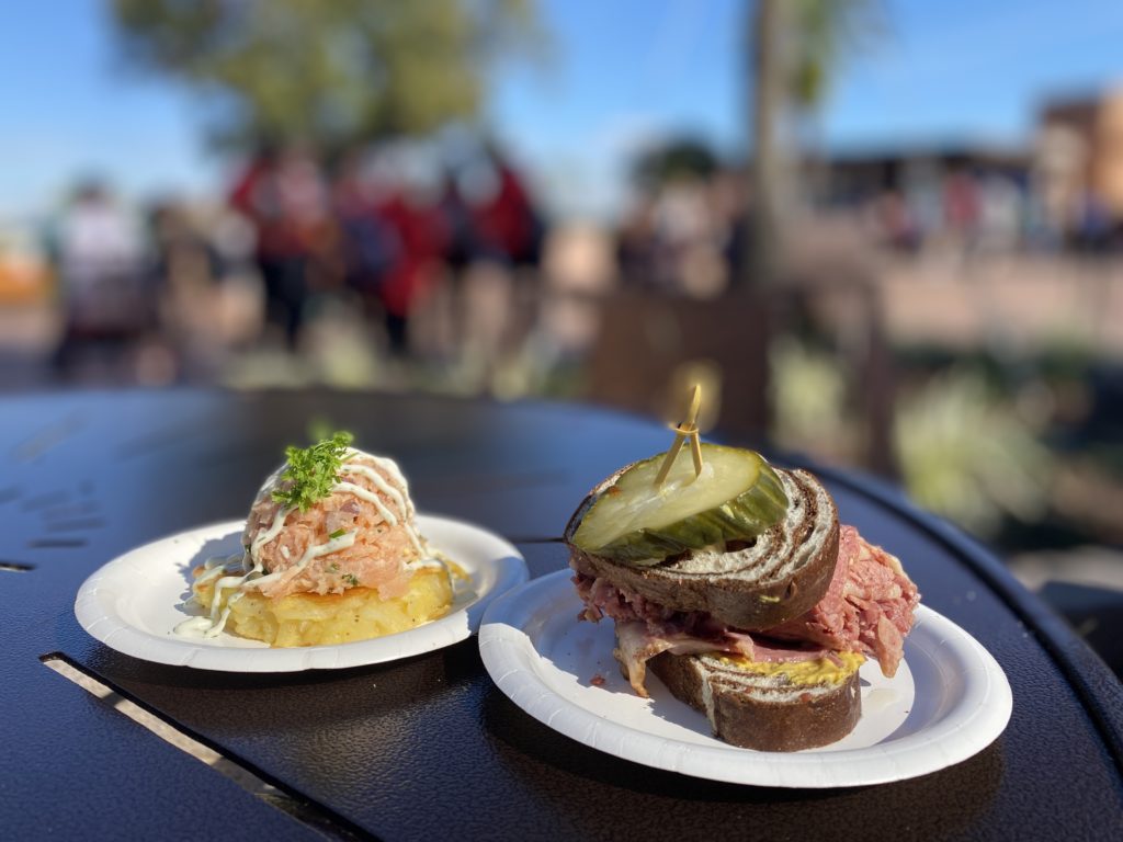Pastrami on Rye and Smoked Salmon Potato Latke - L’Chaim! Holiday Kitchen are served on small white paper plates and placed on a black table. The World Showcase promenade is in the background