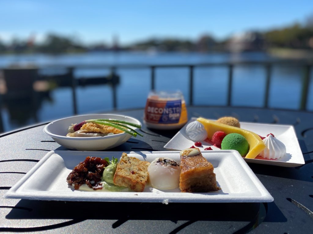 Delicious Food from Deconstructed Dish at 2022 Epcot Festival of the Arts - three white paper plates of artistic looking food sit in the foreground on a black metal table, and a multiple colored short round cup with the Deconstructed Dish logo is in the back of the table, and a blue World Showcase lagoon is blurred in the background