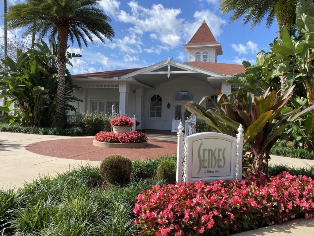 Sense Spa at Disney's Grand Floridian is a freestanding building with white exterior and a terracotta roof. This photo is taken facing the building with a flower bed and sign for the spa in the foreground and blue skies and green palm trees around the building