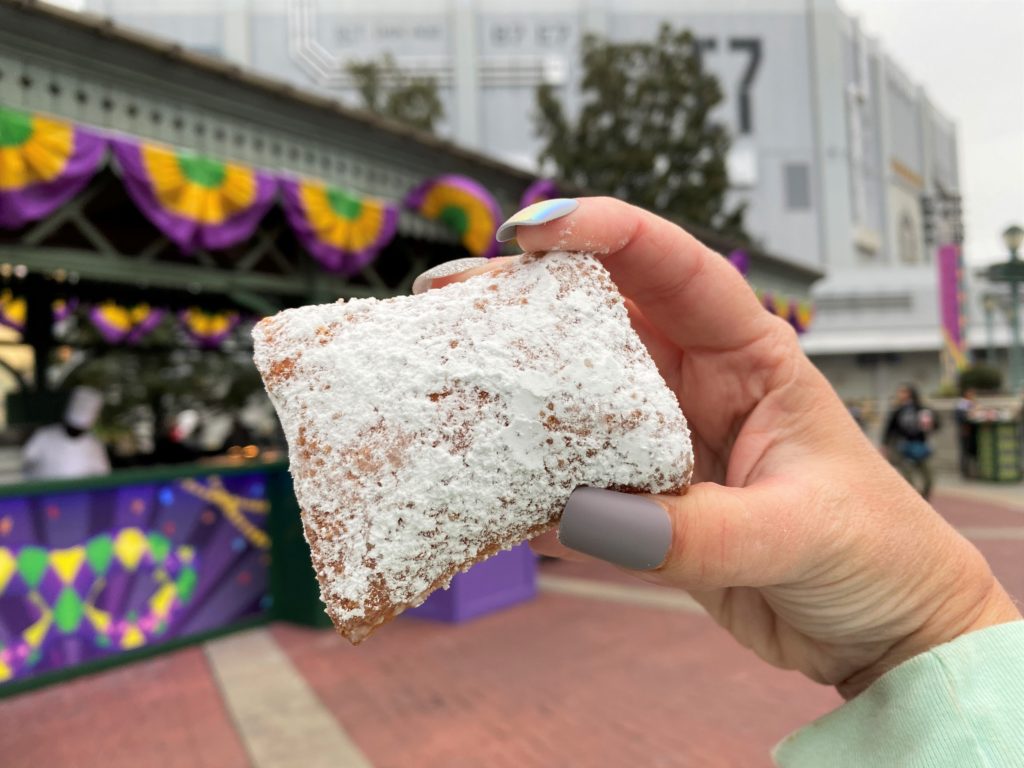 A traditional beignet with powdered sugar is held up in front of a mardi gras decorated food booth